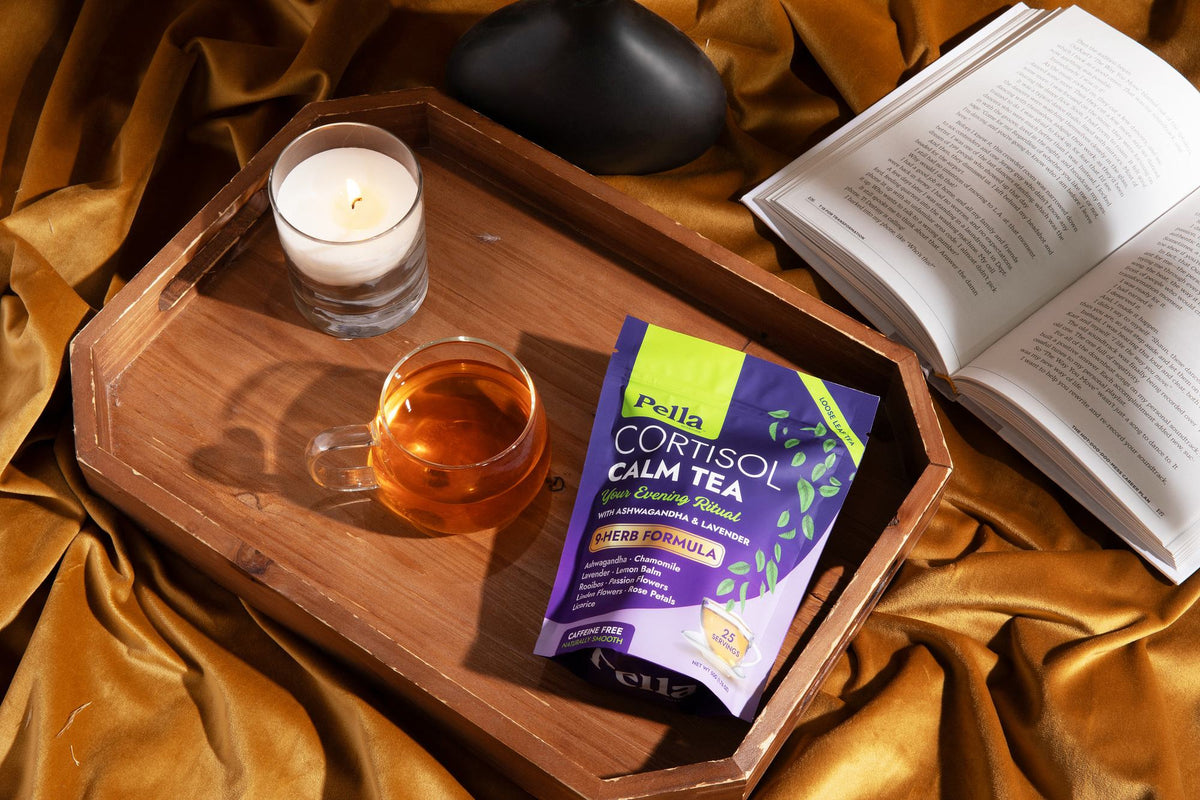 Cortisol Calm Tea pouch on a wooden tray with a glass of brewed tea, a candle and an open book